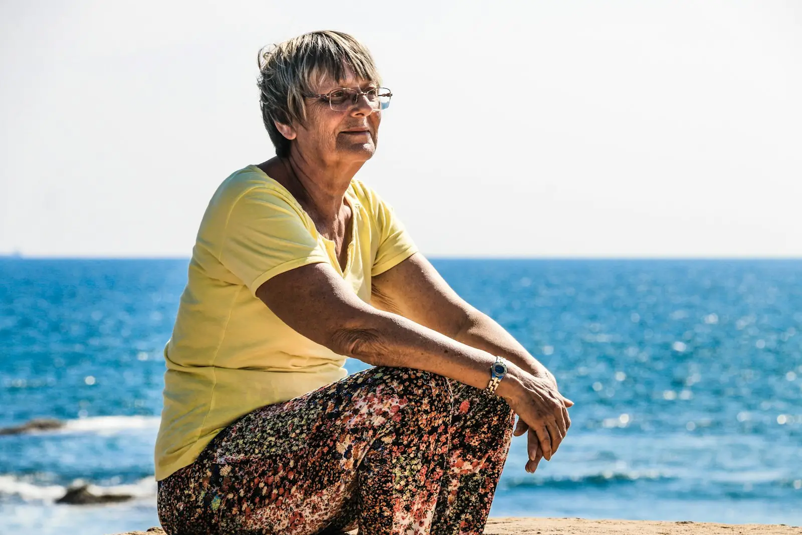 An older woman sitting on rocks by the sea, smiling