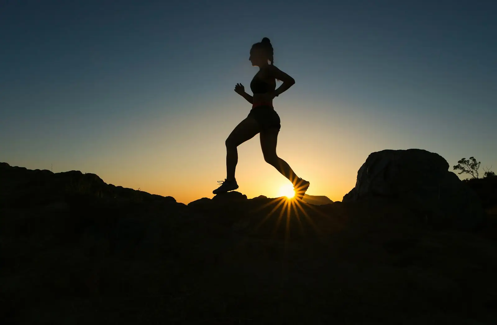 A woman in silhouette running on a rocky trail against a low sun