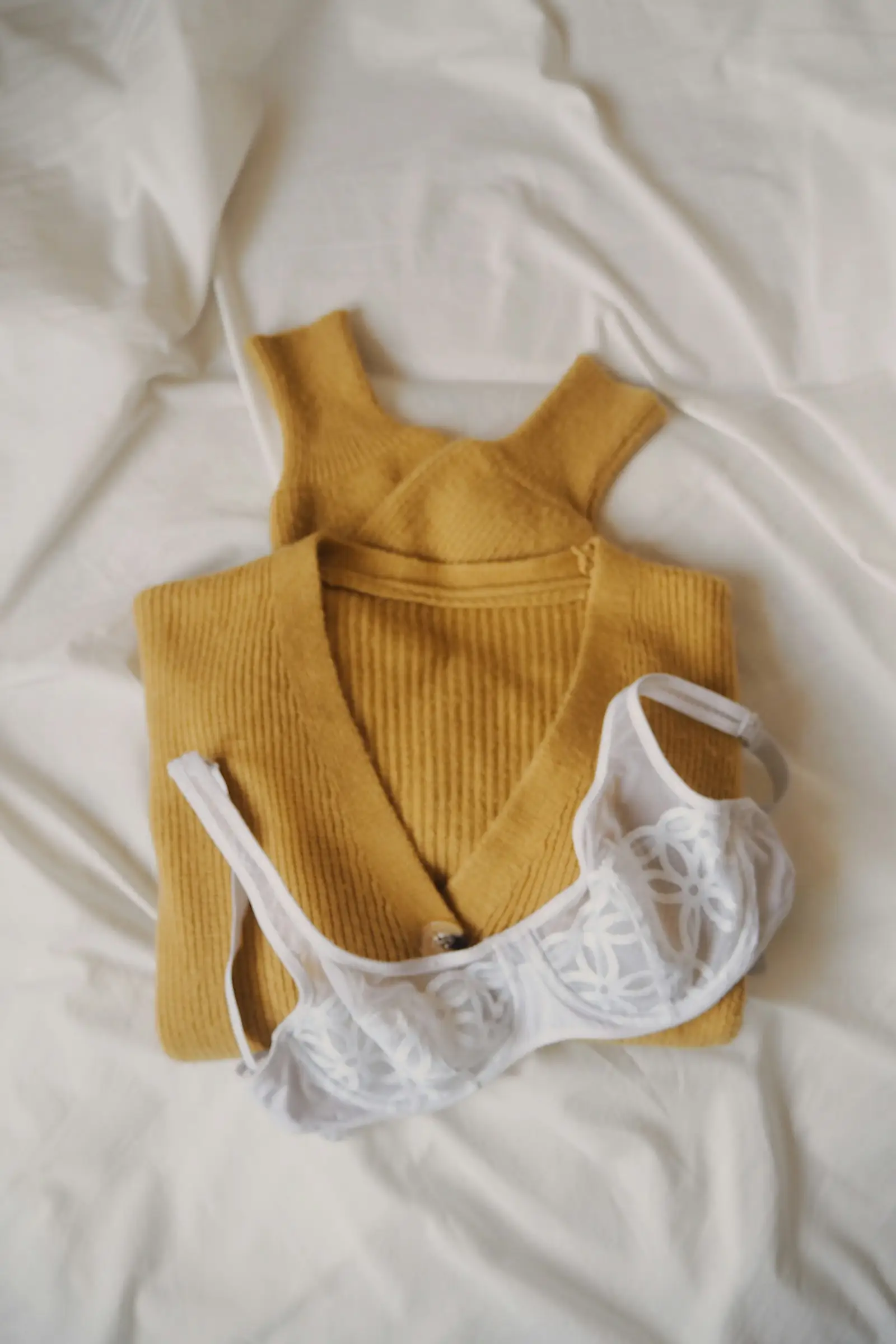 A white lace bra lying on a bed next to a mustard knit top
