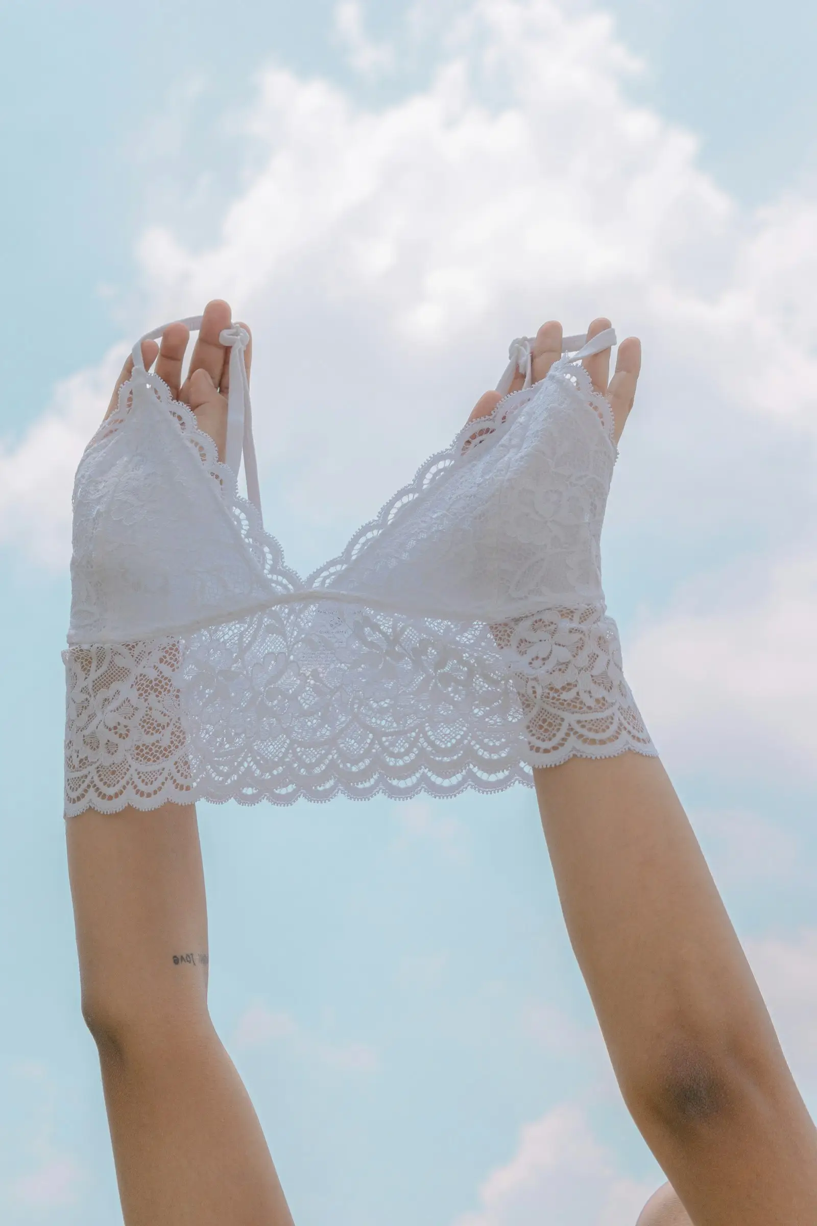 Hands holding up a white lace bralette against a blue sky